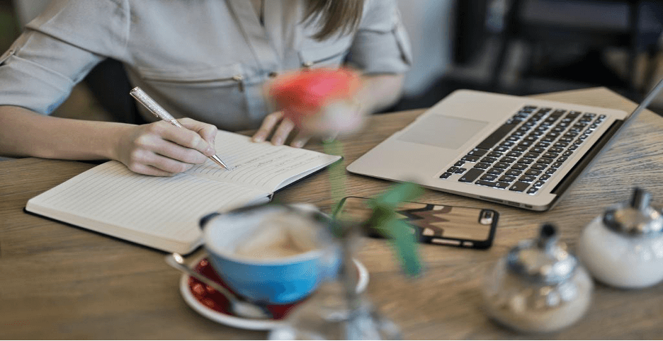 Author writing in a notebook beside a laptop and coffee